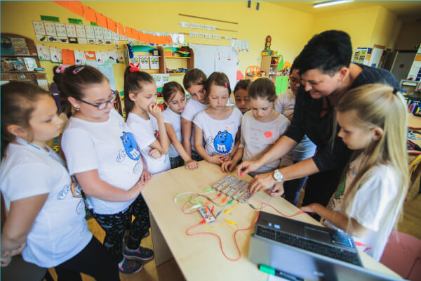 Zdjęcie przedstawia nauczycielkę obsługującą elektronikę tłumacząc ją dzieciom.